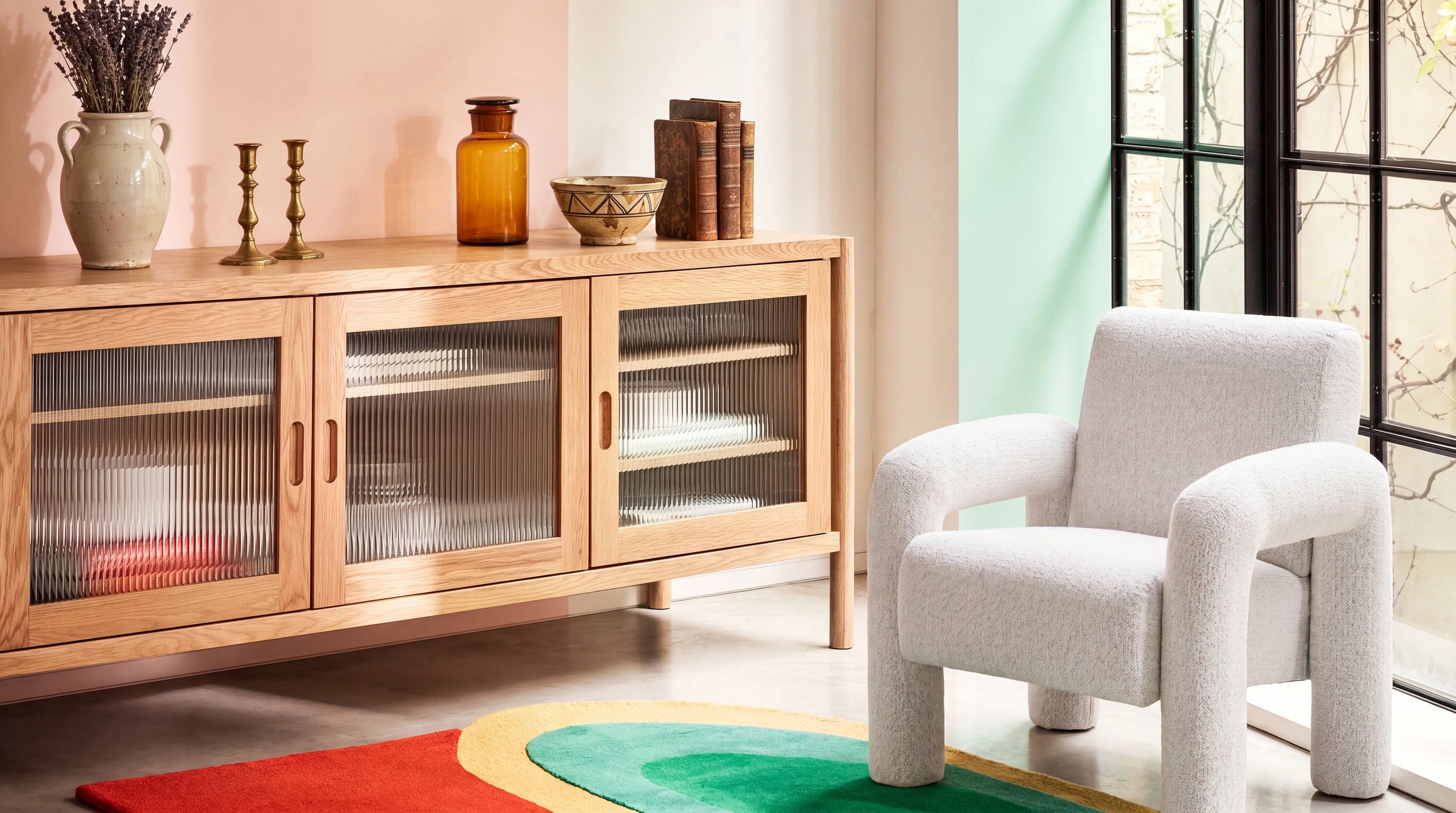 Wooden sideboard with glass doors next to a white armchair in a room with a large window.